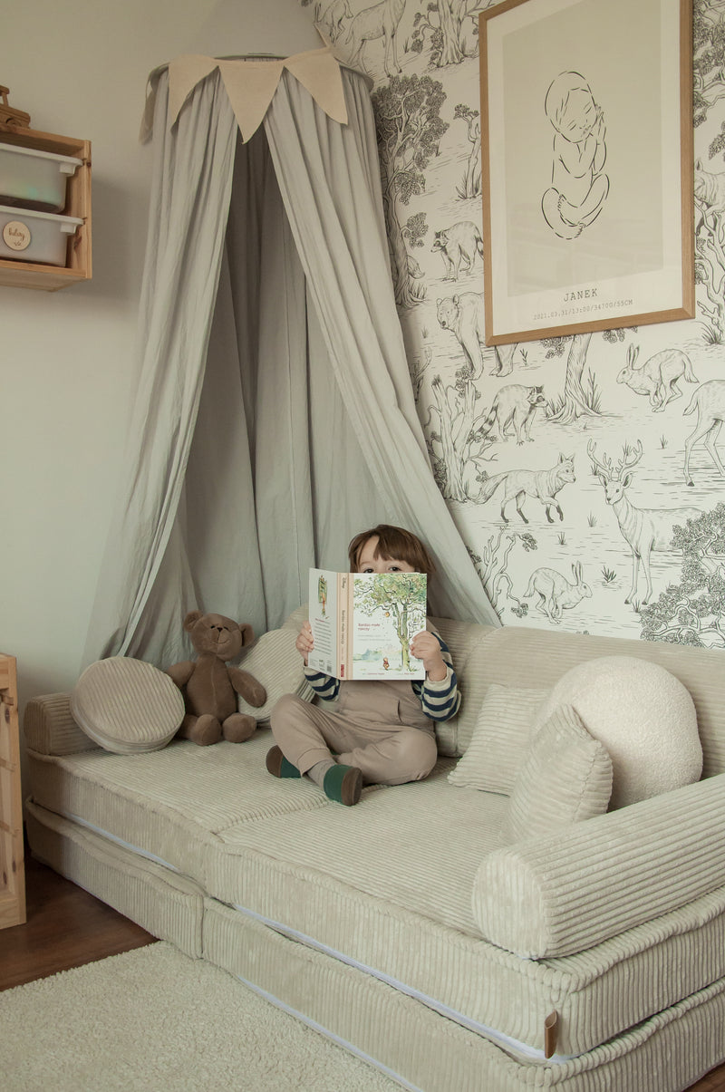 Child reading a book on a bed with a teddy bear in a room with patterned wallpaper.