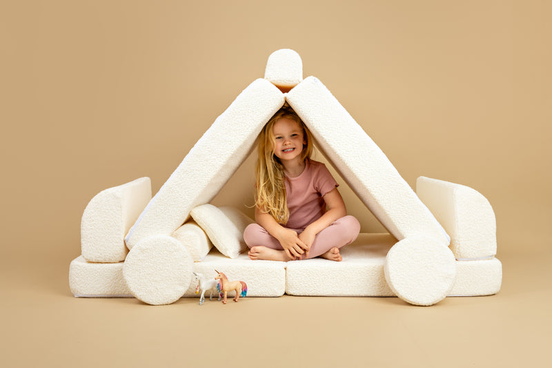 Child sitting inside a pyramid-shaped structure made of white cushions on a beige background